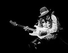 A black and white photograph of Jimi Hendrix playing a Fender Stratocaster electric guitar (during his "hat phase" in 1968).