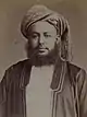 A black-and-white photograph of a man with a dark beard wearing a turban, a dark jacket, a shirt, and a belt, sitting in a chair, and looking at the viewer