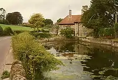 Water contained within stone walls to the right of road. In the background stone house with red roof.