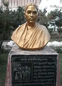 A golden bust of an Indian woman sits atop a concrete pedestal with a large black plaque outside in a park.