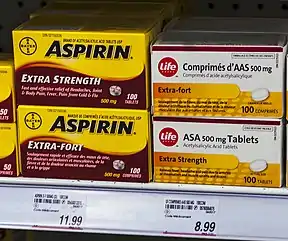 Four boxes of medication on a store shelf above price tags. The two on the left are yellow with "Aspirin" in bold black type and explanatory text in English on the top box and French on the bottom. The two on the right are slightly smaller and white with the word "Life" in the corner inside a red circle. The text, in French on top and English below, describes the medication as "acetylsalicylic acid tablets"