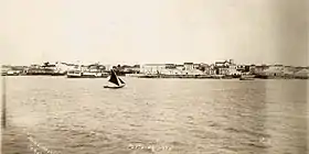 Photograph looking across water with a small sail boat in the foreground and buildings along the shore in the background