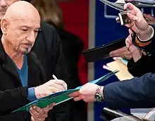 Older man in a blue shirt and black jacket with a beard and moustache signing an autograph