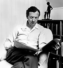 Black and white photograph of a man sitting in a chair; he is holding opened literature