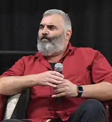 A 46-year-old man with gray hair and a gray beard holding a microphone and looking to the left of the camera.