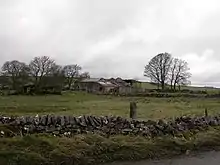 Green farmland on an overcast day, with a stone wall in the foreground, and somewhat dilapidated stone buildings bordered by trees in the background