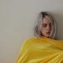 Eilish is seen wearing all yellow, leaning against a wall and looking to the side.