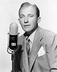 A fair-haired man wearing a light-coloured suit, standing at a microphone