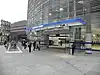 A glass structure with gray slats on higher floor; an entrance leads under a canopy with a sign reading "BLACKFRIARS STATION" into a large internal space seen through the glass, People are walking into the entrance and coming up steps from an underpass.