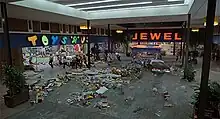 Film screenshot showing a police car driving through a shopping mall: Scattered items are present on the floor and people are running away from the vehicle. Stores visible in the mall include Toys "R" Us and Jewel.