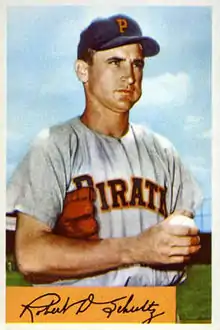 A man in a gray baseball jersey with "Pirates" written on the chest and a navy blue cap with an orange "P" on the front standing with a glove under his arm and a ball in his hand