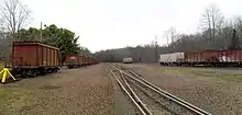 A view of a small rail yard. The main track is in the center and clear, while on several other tracks empty red or white hopper cars are stored. Logos for Tilcon and the reporting marks "TILX" are visible on several of the hoppers.