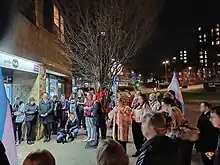 A group of people gathered in a circle on a sidewalk at night. Many are holding transgender pride flags.