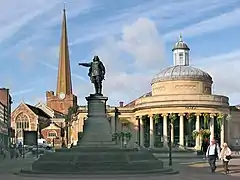 Statue of figure with outsretched arm. To the left a tall church spire and the right a circular building with columns.