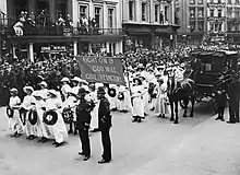 A procession of Suffragettes, dressed in white and bearing wreaths and a banner reading "Fight on and God will give the victory" during the funeral procession of Emily Davison in Morpeth, Northumberland, 13 June 1913. Crowds line the street to watch.