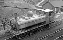 A pannier tank locomotive, seen from above and to the front, is passing through hilly countryside. The locomotive, particularly at the front, is streaked with vertical stains. The lettering "NCB 7754" is shown on the side of the pannier tank.