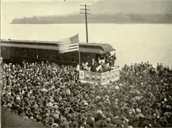 A dramatic political scene. Beside a river stands a podium, on which a flagpole flies a huge American flag. Beneath the flag stands a candidate in a dark suit addressing an impressive crowd which takes up most of the photograph. Not only the quayside but a ferry beside it on the water are packed full of people listening intently.