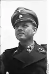 A man wearing a black military uniform, peaked cap, and a neck order in the shape of a cross. His cap has an emblem in shape of a human skull and crossed bones.