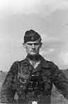 A black-and-white photograph of a man wearing a camouflage military uniform, side cap, and a pair of binoculars around his neck. His cap has an emblem in shape of a human skull and crossed bones.