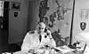 Black-and-white photo of a man wearing a white military uniform sitting at a desk, making a phone call.