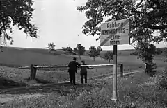 Two people standing either side of a lowered border pole on a dirt road with a sign in the foreground