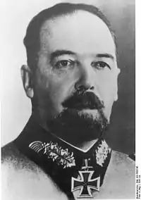 The head of a man with beard, shown in semi-profile. He wears a military uniform with a military decoration in shape of an iron cross displayed at the front of his shirt collar. His hair is dark and combed to back.