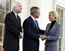 White-haired man in dark suit looks on as gray-haired man in dark suit holds hand and greets blonde-haired woman in medium-colored suit, all in front of a white building.