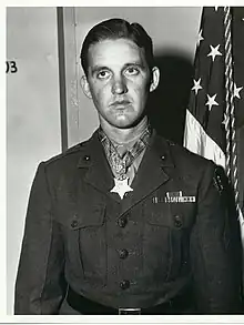 Head and torso of a young white man with neatly combed hair wearing a dark military jacket with a wide belt and a few ribbon bars on the left breast. A star-shaped medal hangs from a ribbon around his neck.