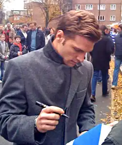 A man wearing a dark grey woollen coat signs an autograph outside for a fan.