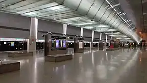 A large island platform which include flight information displays. An illuminated linkbridge spans over the platform.