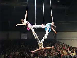 Two children hang from a suspended bar, supporting a third child with their feet. The contours of their legs form a triangle shape.