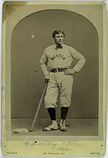 A man, wearing a baseball uniform with the words "BOSTON" in the center, leans on a bat held in his right hand.