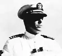 Head and shoulders of a man in a white jacket with black shoulderboards with binoculars hanging from around his neck. His eyes are shaded by a white peaked cap with a black visor.