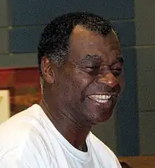 A headshot of a black man. He is looking to the right, wearing a plain white T-shirt and is smiling.