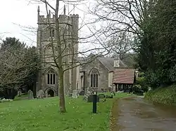 Square grey tower of stone church building, partially obscured by trees. Red roofed lych gate to right. Grass and gravestones in the foreground