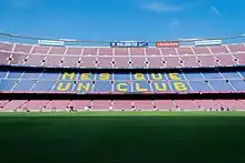 The words "Més que un club" are painted in yellow on the blue seats of the stadium