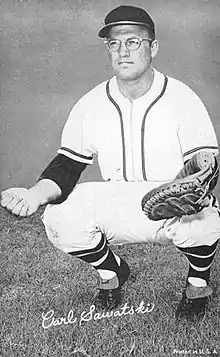 A man in a light baseball uniform, dark cap, and eye glasses squatting in preparation to catch a ball in his mitt