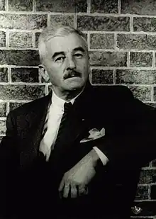 A photograph of William Faulkner in a suit and with a mustached, reclined against a brick wall