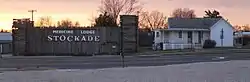 Log stockade with low square towers at corners, bearing large sign "Medicine Lodge Stockade", next to one-story frame house with wraparound porch