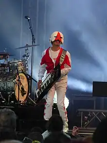 White male standing on musical stage holding a keyboard, wearing red-and-white-patterned pants, jacket, and matching face-shielding helmet, smiling at audience.