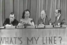 Three men and one woman sit at a long table. Behind them is a curtain. On the front of the table are the words "What's My Line?".