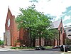 An L-shaped brick building with two pointed roofs and a large tree in its crook