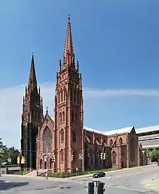 A church of ornate brown stone with two tall towers in front seen from across an intersection.