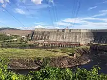 a river flows smoothly from rectangular openings at the base of a high sloping concrete wall, with electricity wires above the river