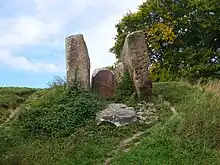 Three large stone slabs forming a chamber, as seen from below. The sky behind is white, and the stones are surrounded by green grass.