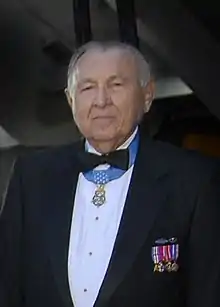 Head and shoulders of an elderly white man with a round face, wearing a tuxedo with military medals pinned to the chest and one medal hanging from a light blue ribbon around his neck.