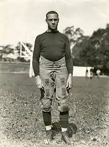 An African American male in a 1920s football uniform stands on a football field. Several unidentifiable individuals are visible in the distance