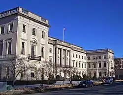 A three-story stone building, seen from its left, with two projecting wings and a balustrade running along the top. An American flag flies from a flagpole above the main entrance at the center, where a row of four columns marks the main entrance. There is an iron fence in front and small iron balconies on the wings.