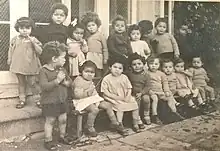 8 17 mixed race children sitting and standing outside the Holnicote House building in England in the 1940s. The children are wearing dresses or shorts.