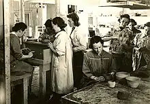 In a ceramics studio, several students stand as they watch their instructor work, seated, at a pottery wheel.